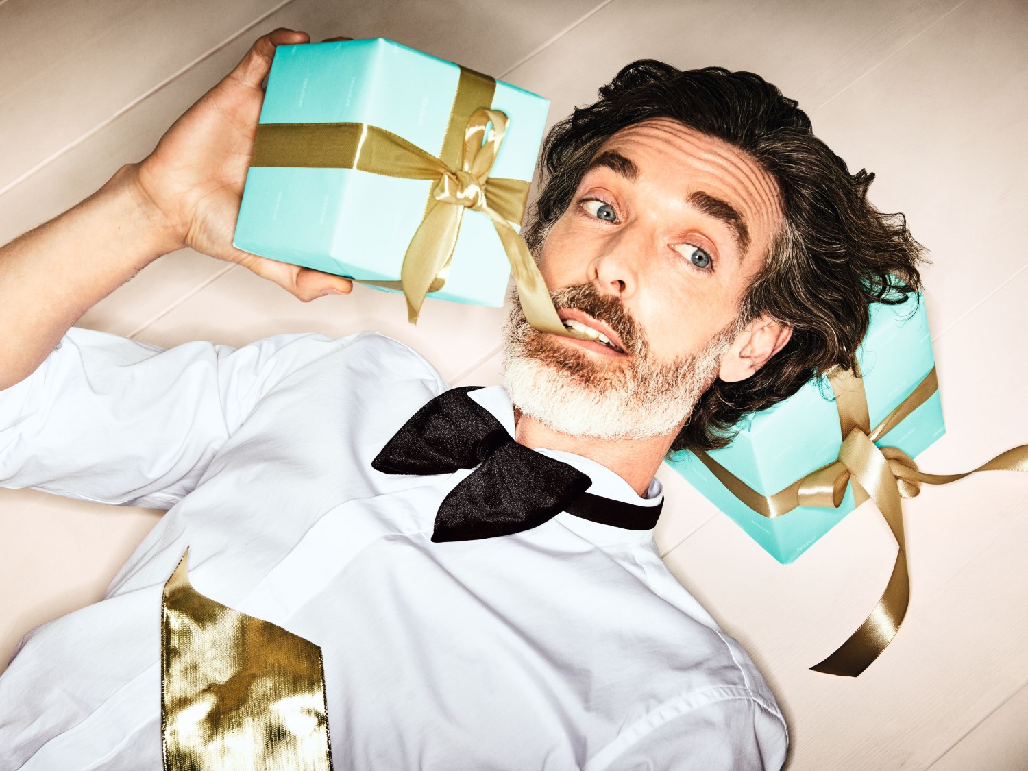 Muž s bradou a motýlikom leží na zemi s dvoma svetlomodrými darčekovými krabičkami so zlatými stuhami.