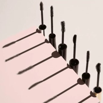 Sześć czarnych maskar do rzęs z aplikatorami ułożonych na różowym tle, rzucających długie cienie.