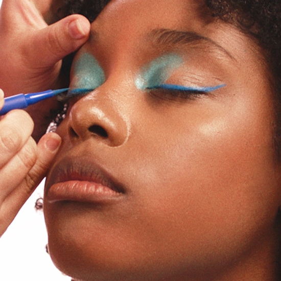 Eine Hand trägt mit einem blauen Eyeliner einen Lidstrich auf das obere Augenlid eines Models auf, das hellblauen Lidschatten trägt.