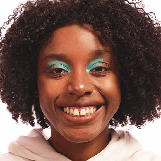 Nahaufnahme einer lächelnden Frau mit lockigem Haar und auffälligem, türkisfarbenem Lidschatten.