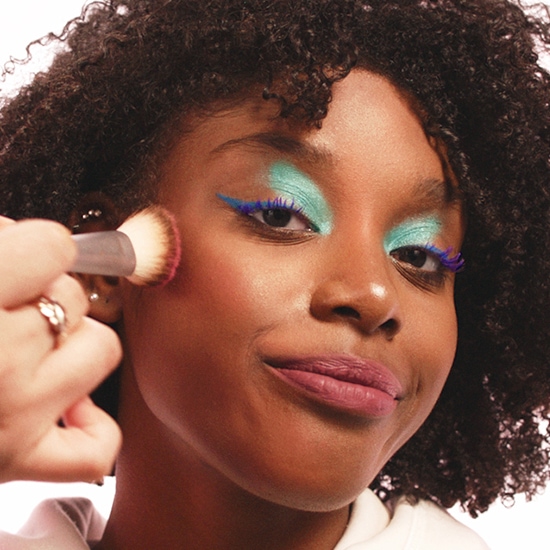 Nahaufnahme einer Frau, die helltürkisen Lidschatten, blauen Eyeliner und lila Wimperntusche trägt, während ein Pinsel Rouge auf ihre Wange aufträgt.