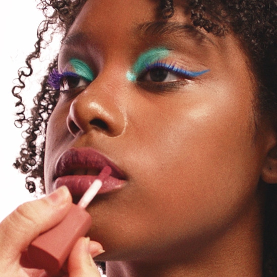 Nahaufnahme einer Frau mit lebhaftem Make-up, einschließlich blauem Eyeliner, türkisfarbenem Lidschatten und violetten Wimpern, die mit einem Pinsel einen glänzenden Lippenstift aufträgt.