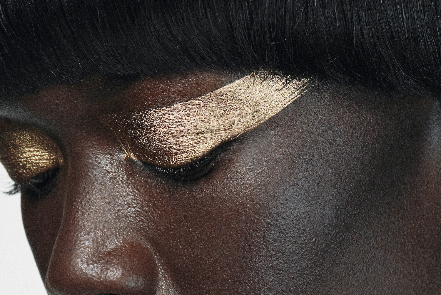 Primer plano del rostro de una mujer de piel oscura con sombra de ojos dorada brillante, que se extiende desde el párpado hasta la ceja, con cabello negro corto y liso.