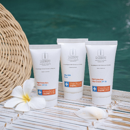 Trzy białe tubki kremów MBR Medical Suncare: High Protection Cream SPF 50, After Sun Face i High Protection Face Cream SPF 30, leżą obok białego kwiatu i muszelki na tle basenu.