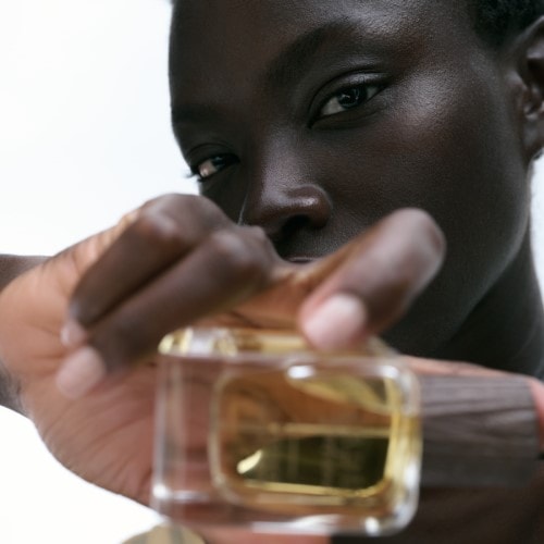 Kobieta trzyma w dłoni flakon perfum o przezroczystych ściankach i złotej zawartości, prezentując go z bliska.