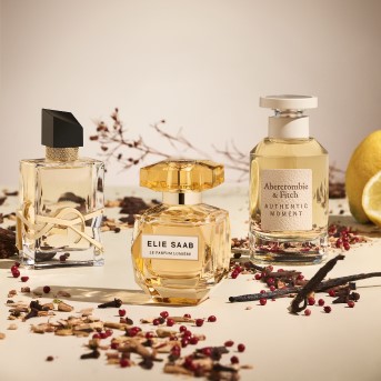 Trzy flakony perfum: YSL z czarnym korkiem, Elie Saab w złotym flakonie i Abercrombie & Fitch Authentic Moment, otoczone przyprawami i cytryną.