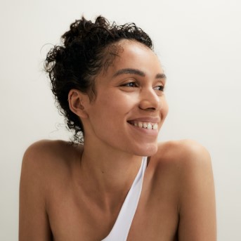 Nahaufnahme einer lächelnden Frau mit lockigem Haar und natürlichem Hautbild, die ein weißes Oberteil trägt.