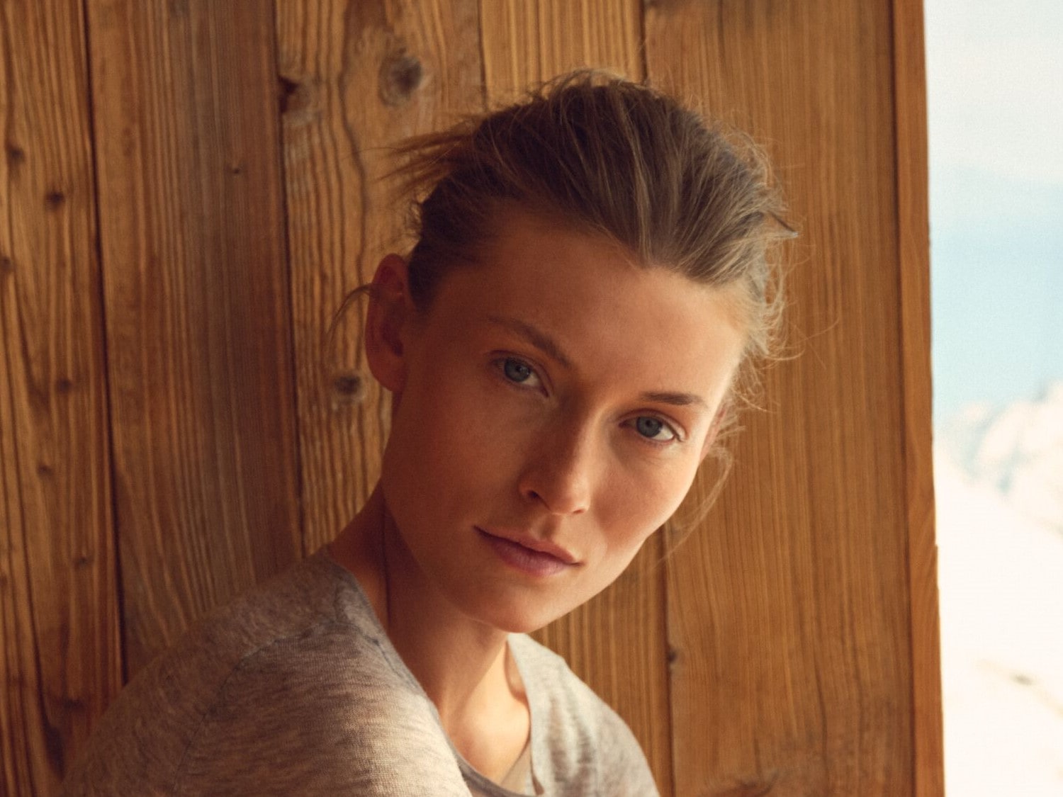 Nahaufnahme einer Frau mit blonden Haaren, die ein graues Oberteil trägt und vor einem Holzhintergrund mit einem Fenster im Hintergrund steht.