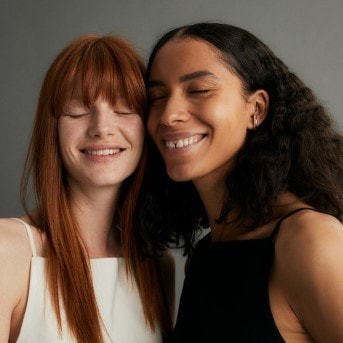 Zwei strahlende Frauen, eine mit roten Haaren und weißem Oberteil, die andere mit dunklen Haaren und schwarzem Oberteil, lächeln mit geschlossenen Augen.