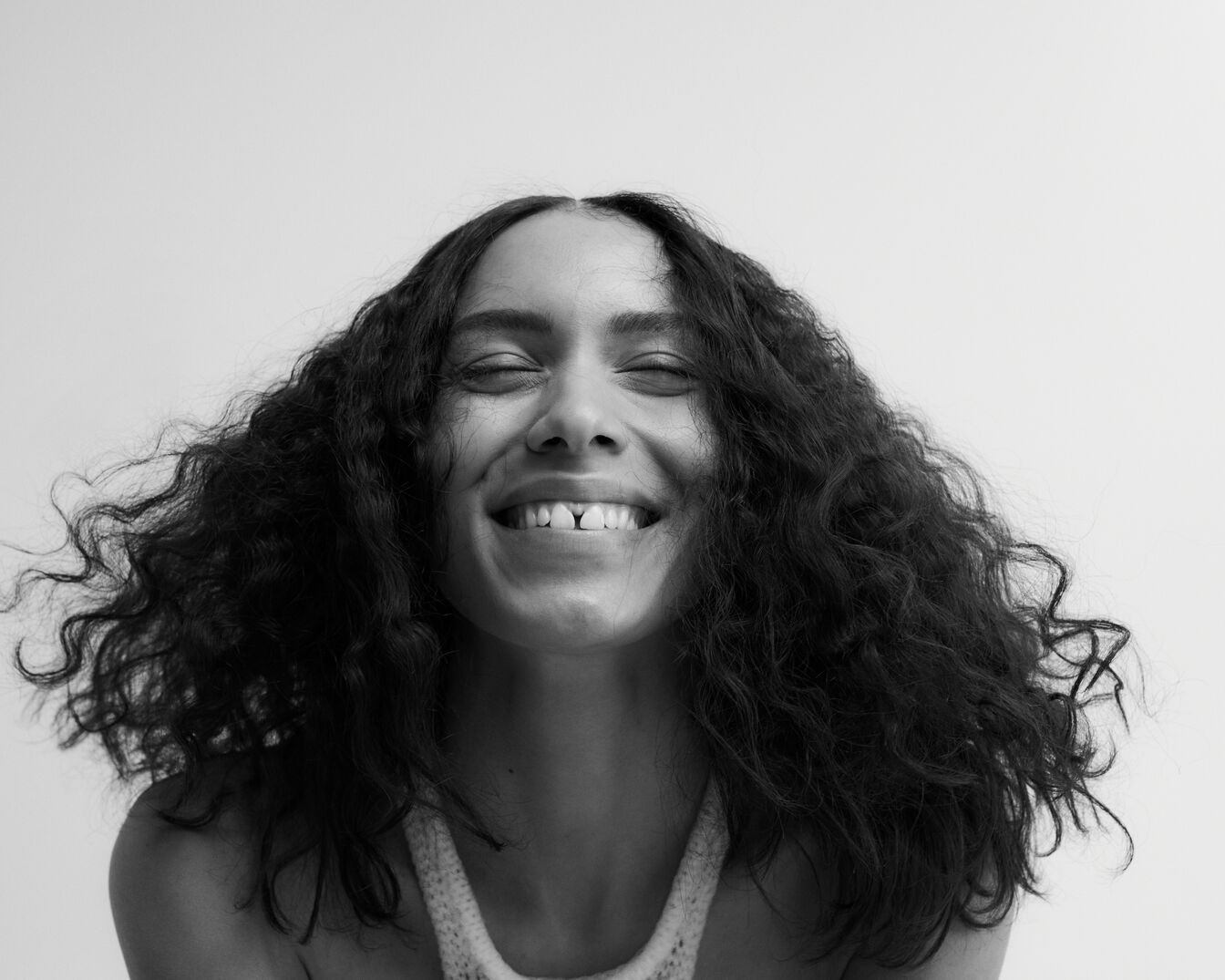 En primer plano, una mujer sonriente con cabello rizado y oscuro, inclinando la cabeza hacia atrás con los ojos cerrados, irradia alegría en una imagen en blanco y negro.