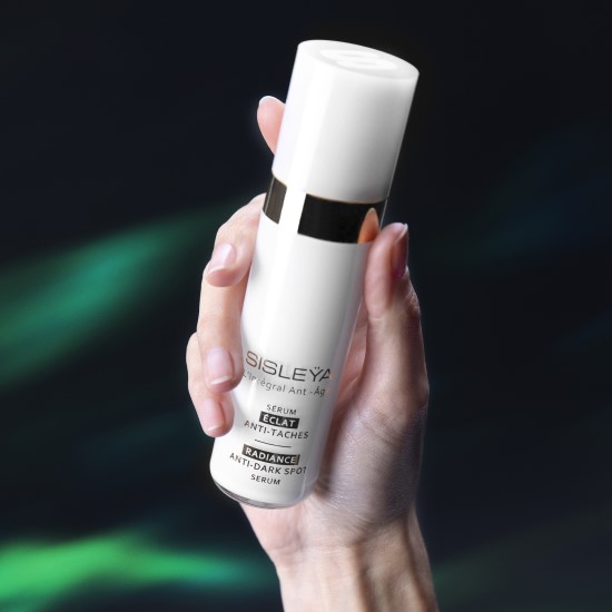 Dłoń trzyma białą butelkę serum Sisleÿa L'Intégral Anti-Âge Radiance Anti-Dark Spot Serum z czarnymi napisami i złotym paskiem.