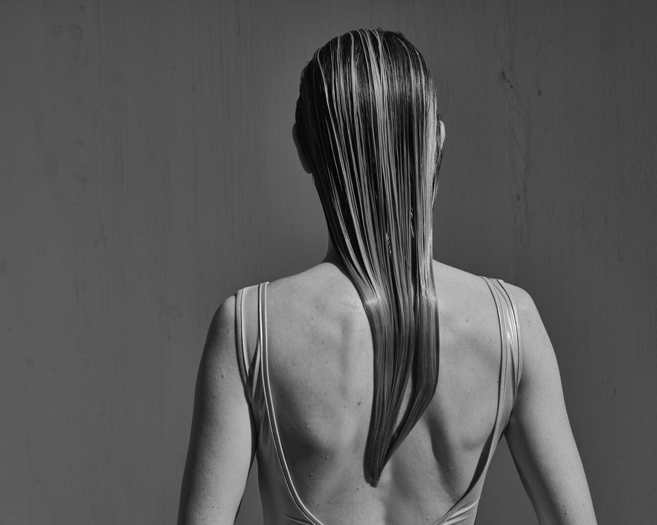 Vista posterior de una mujer con el pelo rubio mojado y liso que le llega hasta la espalda, con un traje de baño de tirantes finos, sobre un fondo gris liso.