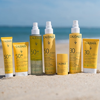 Eine Auswahl an Sonnenschutzprodukten von Caudalie mit LSF 30 und 50+ in gelben Verpackungen, die am Strand stehen.