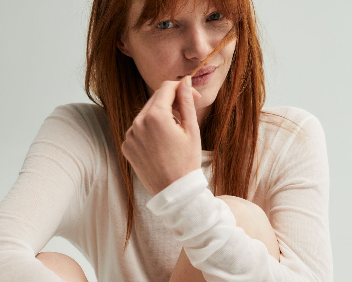 Una mujer pelirroja con una camisa blanca de manga larga sostiene un mechón de su cabello cerca de sus labios, mirando directamente a la cámara con una expresión serena.