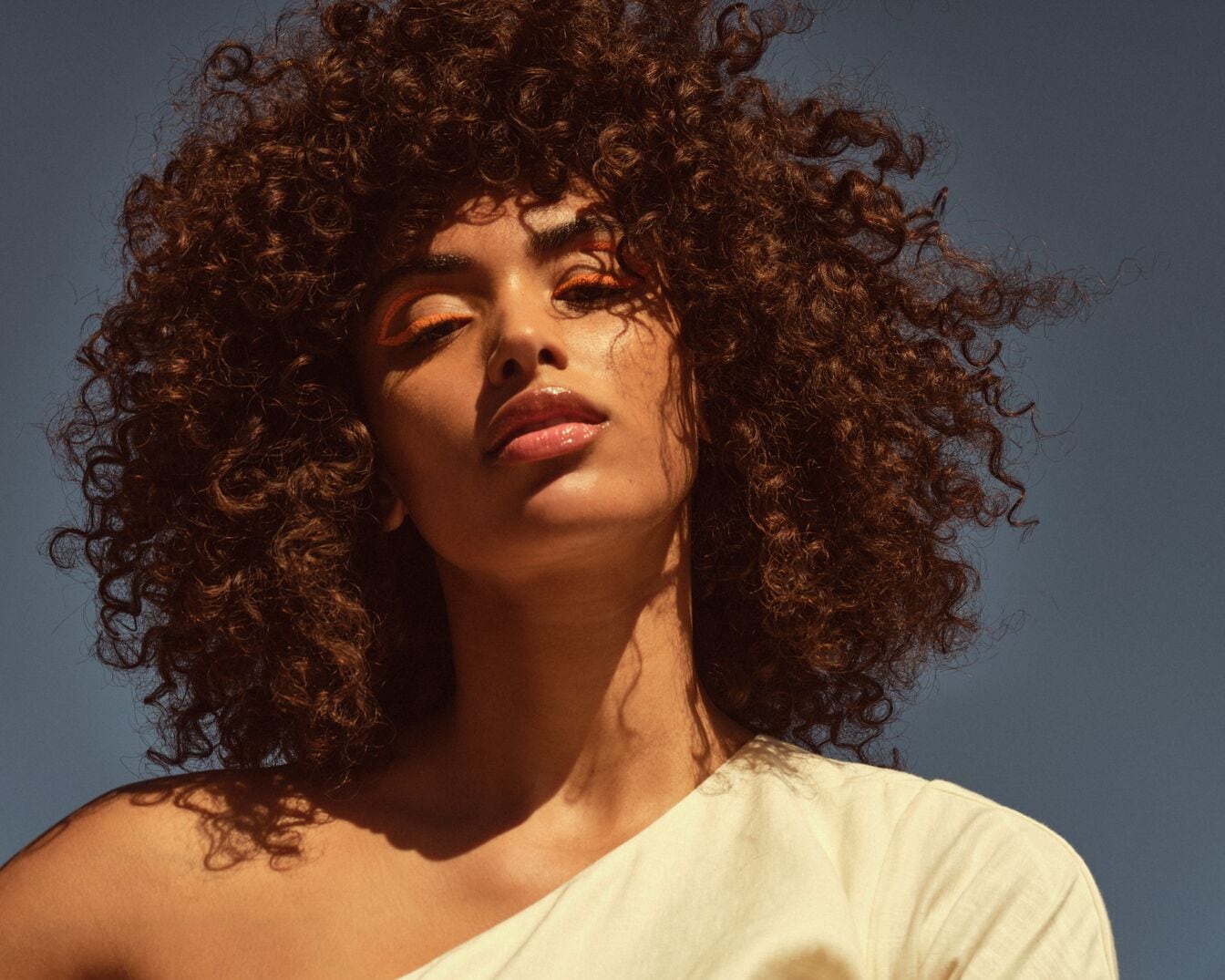 Una mujer de piel morena con cabello rizado castaño y maquillaje de ojos naranja brillante, posa con un top blanco de un solo hombro contra un cielo azul.