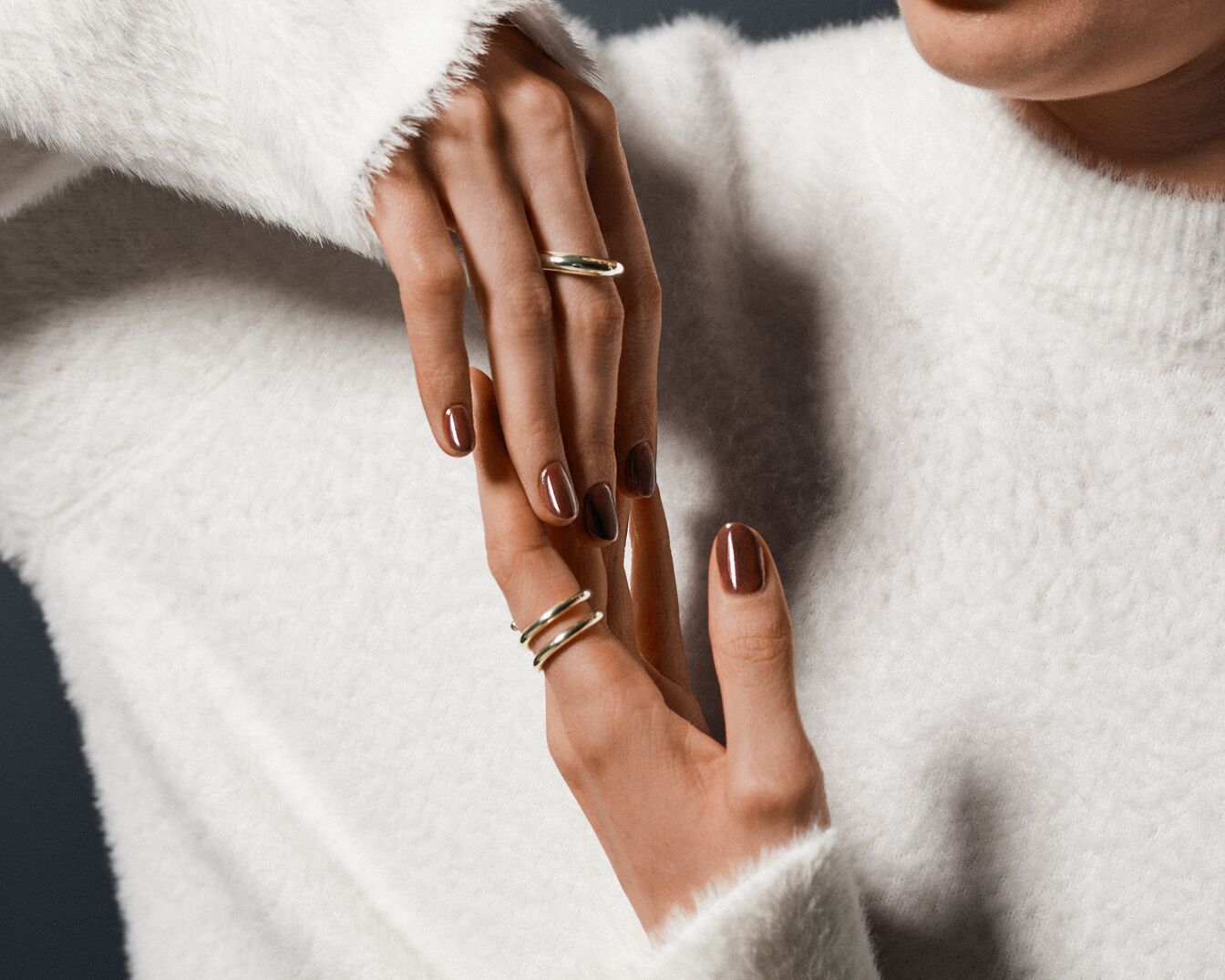 Las manos de una mujer con un suéter blanco y anillos dorados muestran un elegante esmalte de uñas marrón.