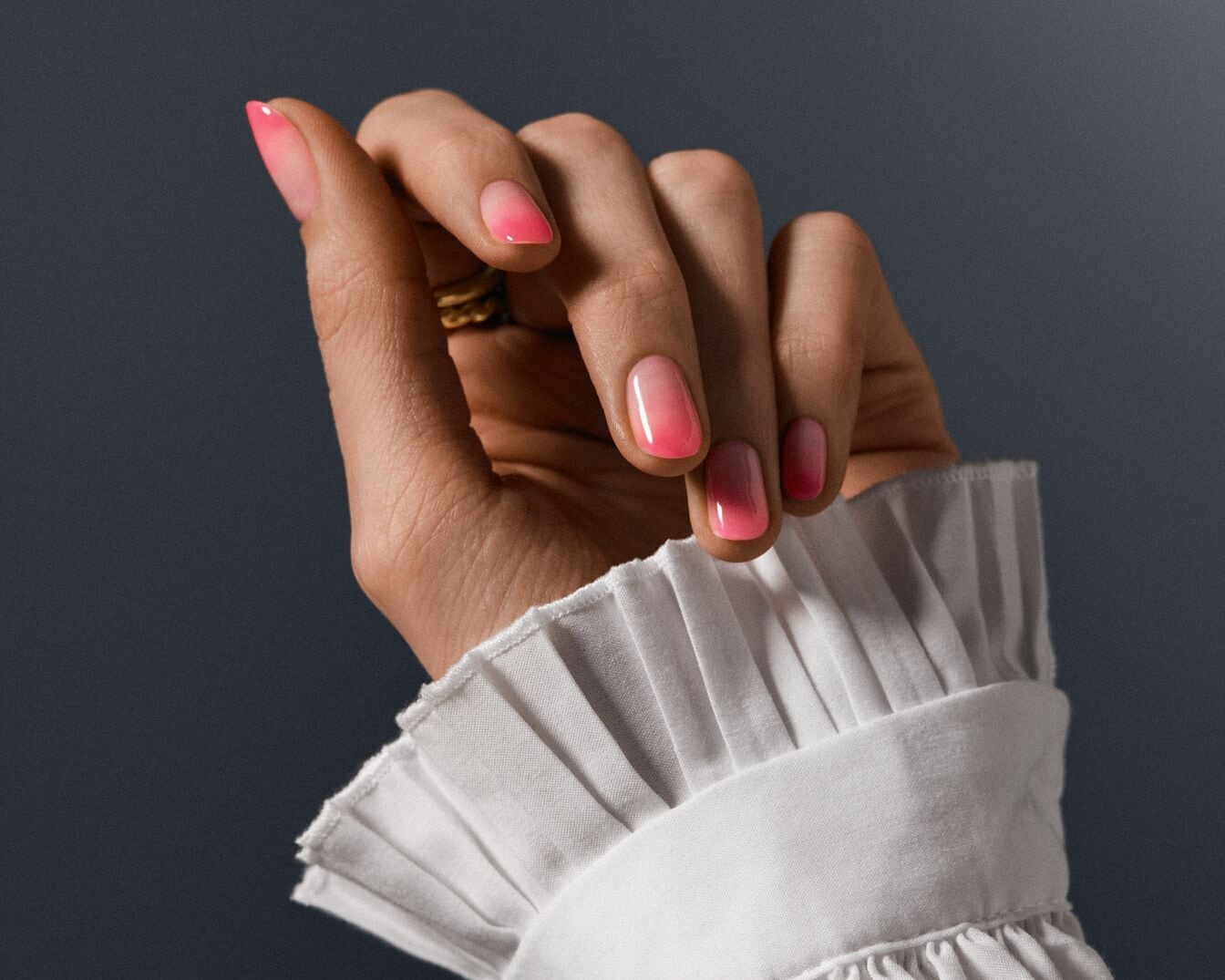 Mano femenina con manicura degradada en tonos rosa y blanco, adornada con un anillo dorado, sobre un fondo grisáceo y un puño de camisa blanco con volantes.