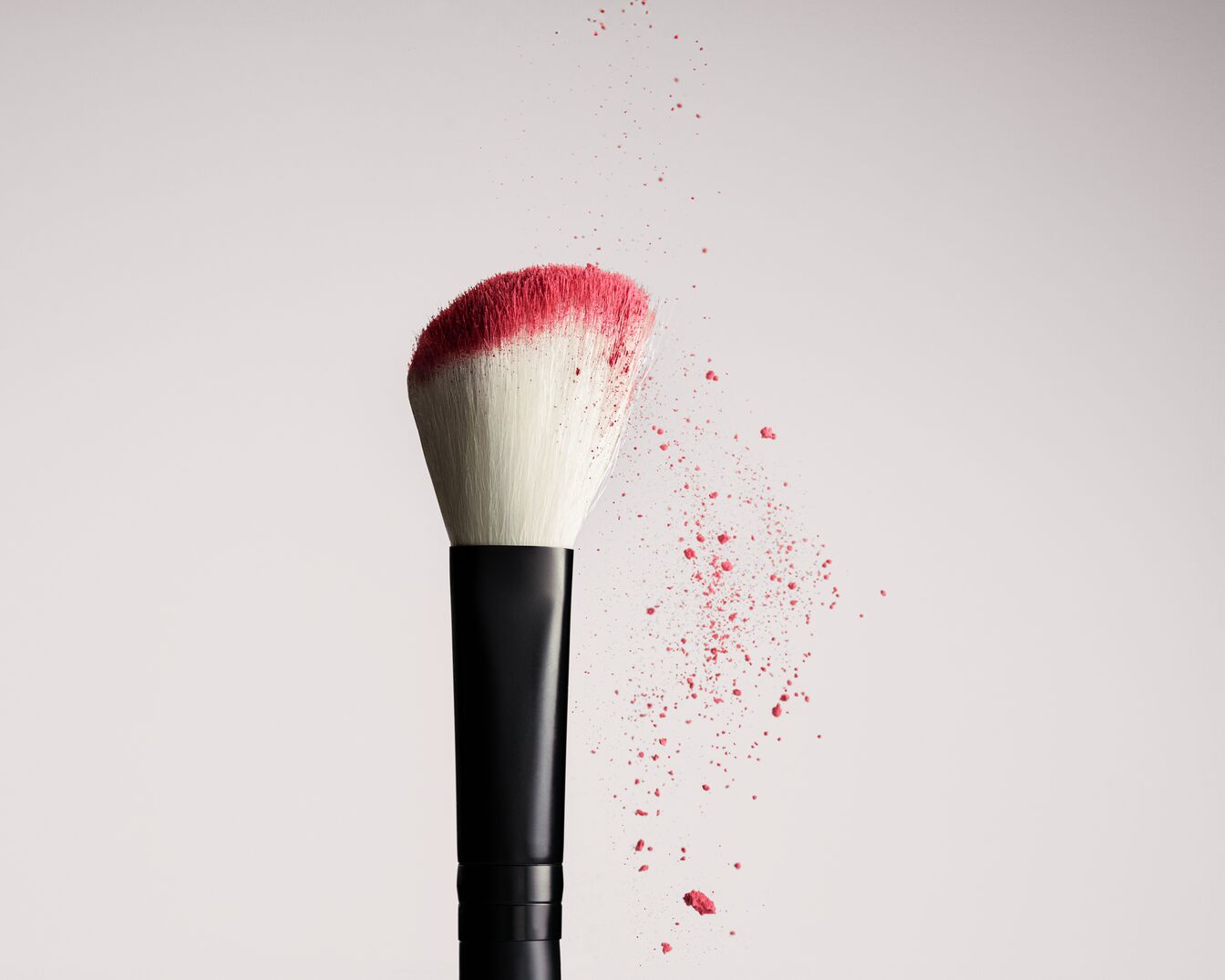 Brocha de maquillaje con mango negro y cerdas blancas, impregnada de polvo rosa que se desprende al aplicarla.