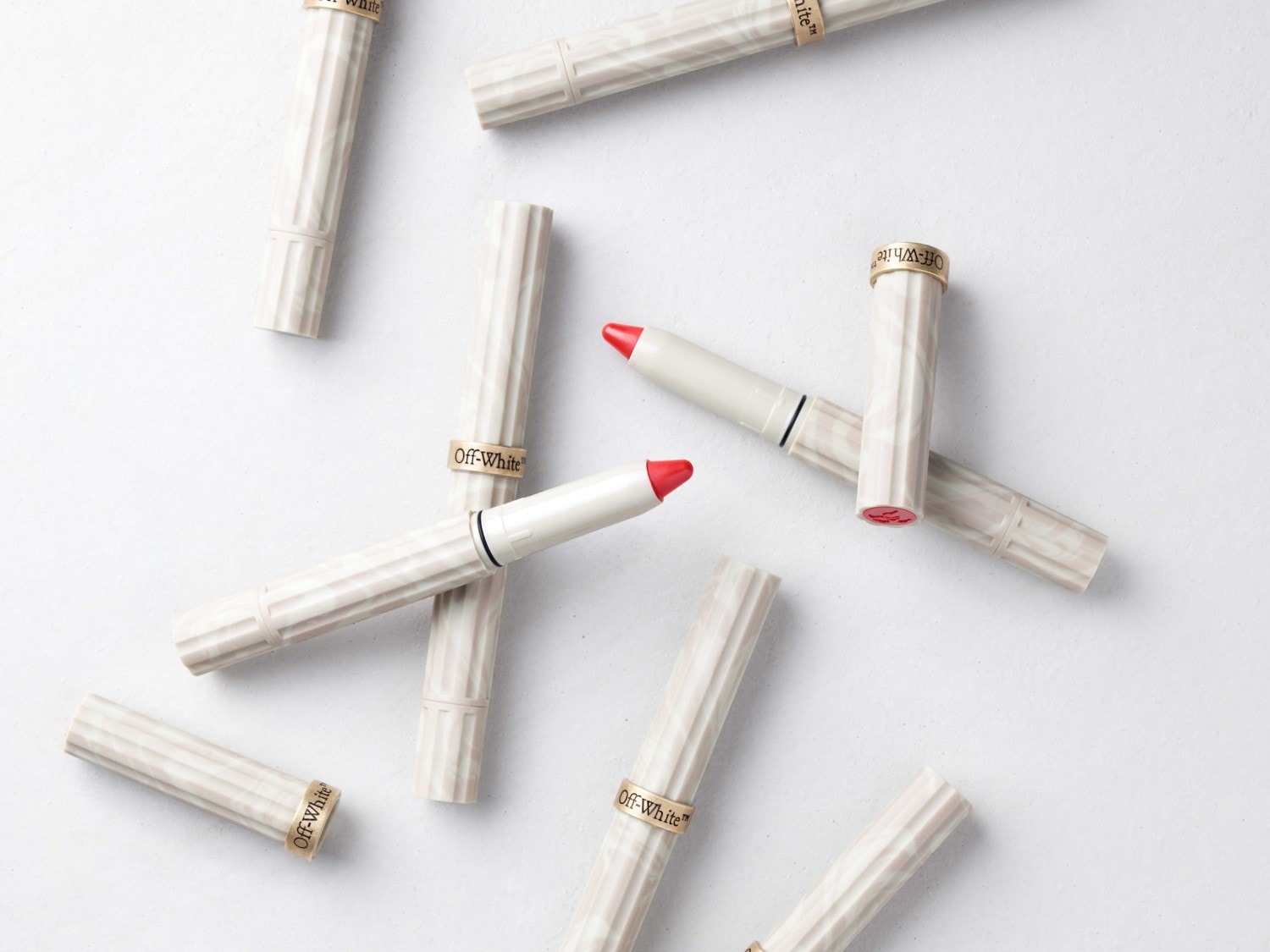 Na białym tle leżą szminki Off-White w beżowych opakowaniach z marmurkowym wzorem i złotym napisem, niektóre z czerwonym wkładem.