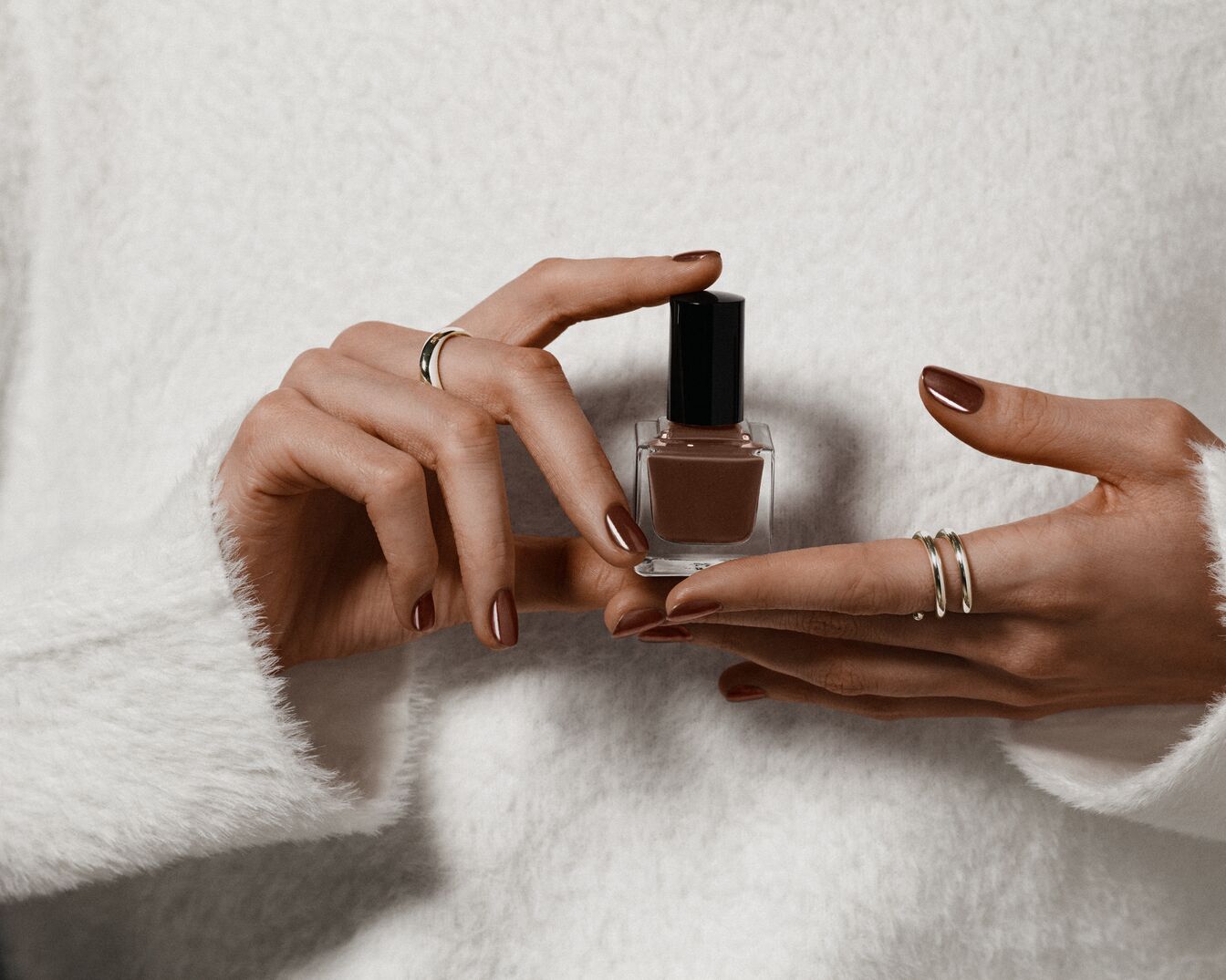 Primer plano de unas manos femeninas sosteniendo un esmalte de uñas marrón chocolate en un frasco de vidrio cuadrado con tapa negra, sobre un fondo de suéter blanco.