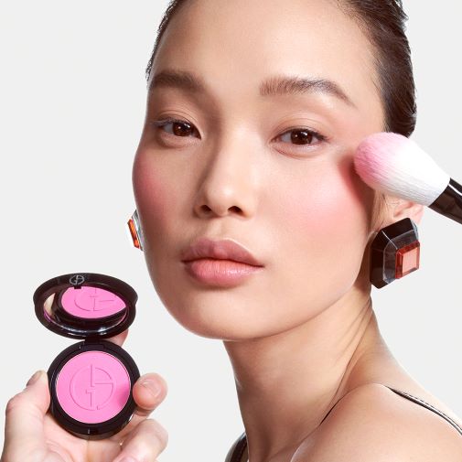 Nahaufnahme eines Models, das rosa Rouge mit einem Pinsel auf die Wange aufträgt, während es eine geöffnete, runde, schwarze Puderdose mit rosa Rouge in der Hand hält.