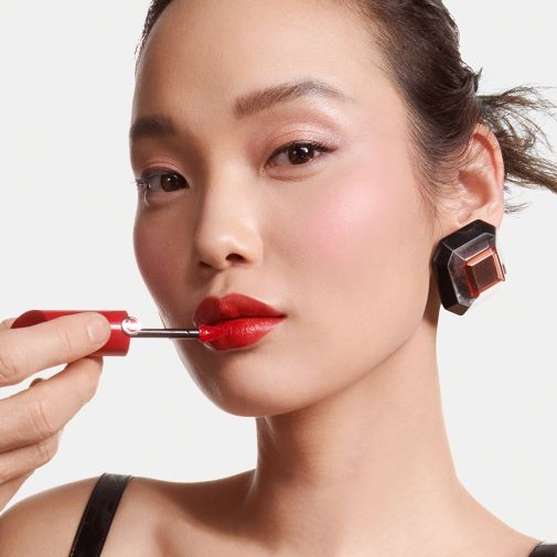 Nahaufnahme einer Frau, die roten Lipgloss mit einem Applikator aufträgt, der in einer roten Hülle steckt, und einen auffälligen Ohrring trägt.