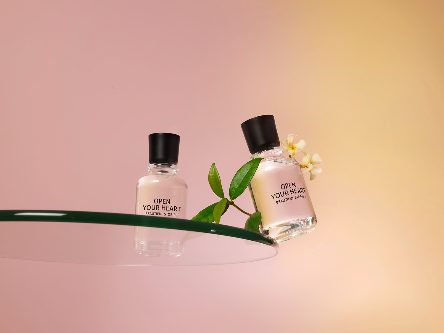 Twee flessen Open Your Heart Beautiful Stories parfum staan op een glazen oppervlak, de ene rechtop en de andere leunend, versierd met een takje en witte bloemen.