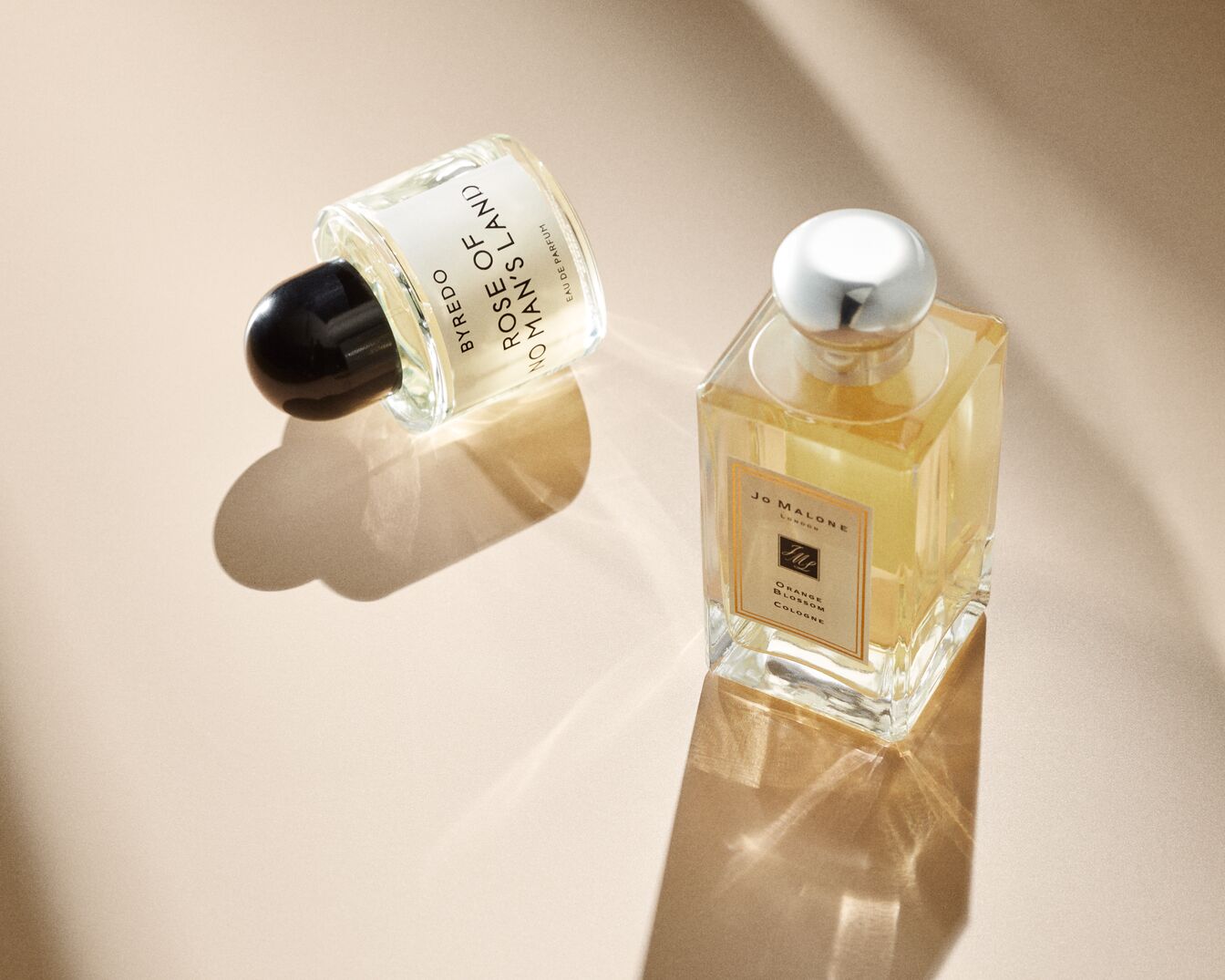 Zwei Parfümflaschen, eine von Byredo Rose of No Man's Land mit schwarzem Verschluss und eine von Jo Malone Orange Blossom Cologne mit silbernem Verschluss, stehen auf einem beigen Untergrund.
