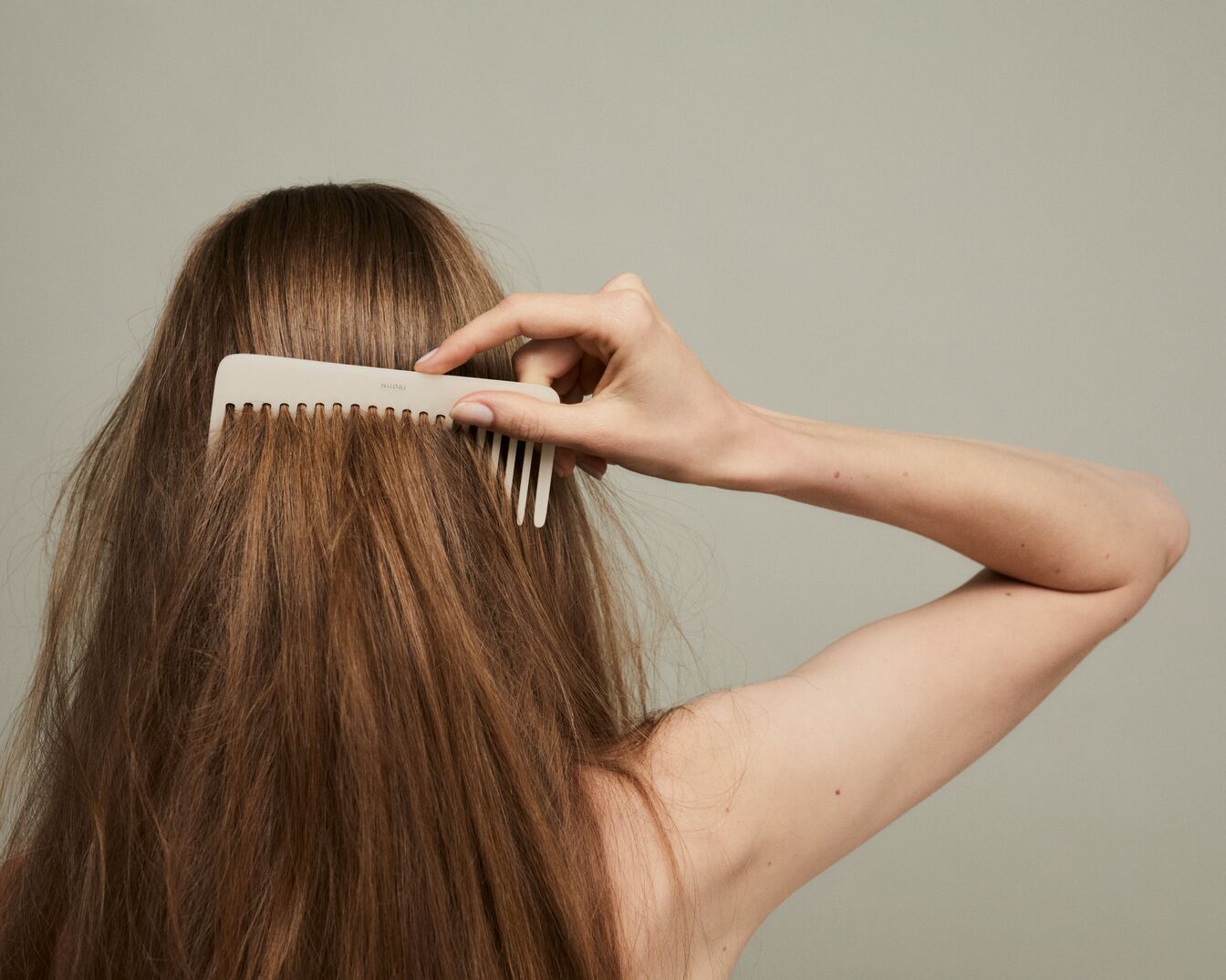 Vista posterior de una mujer peinándose el cabello castaño con un peine color crema.