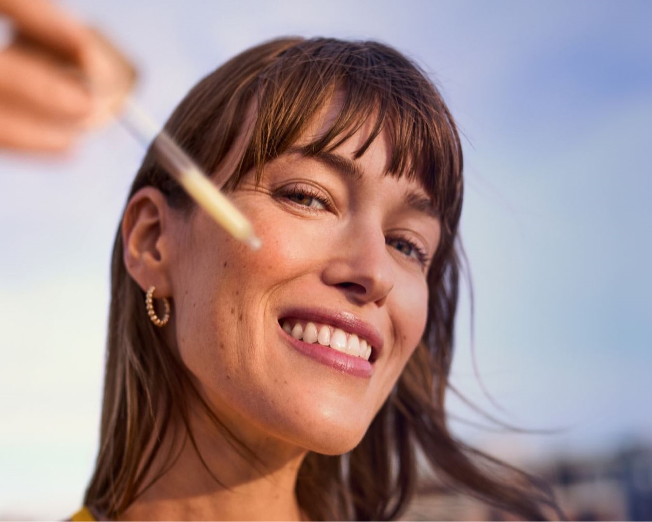 Nahaufnahme einer Frau, die lächelt, während eine Pipette mit Serum auf ihre Wange aufgetragen wird, um ihre Haut zu pflegen.