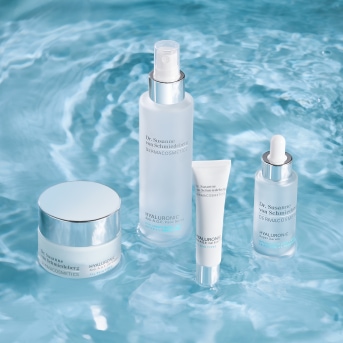 Una gamma di prodotti Dr. Susanne von Schmiedeberg DERMACOSMETICS Hyaluronic, tra cui crema, spray, tubo e siero, tutti confezionati in bianco e argento su uno sfondo blu acqua.