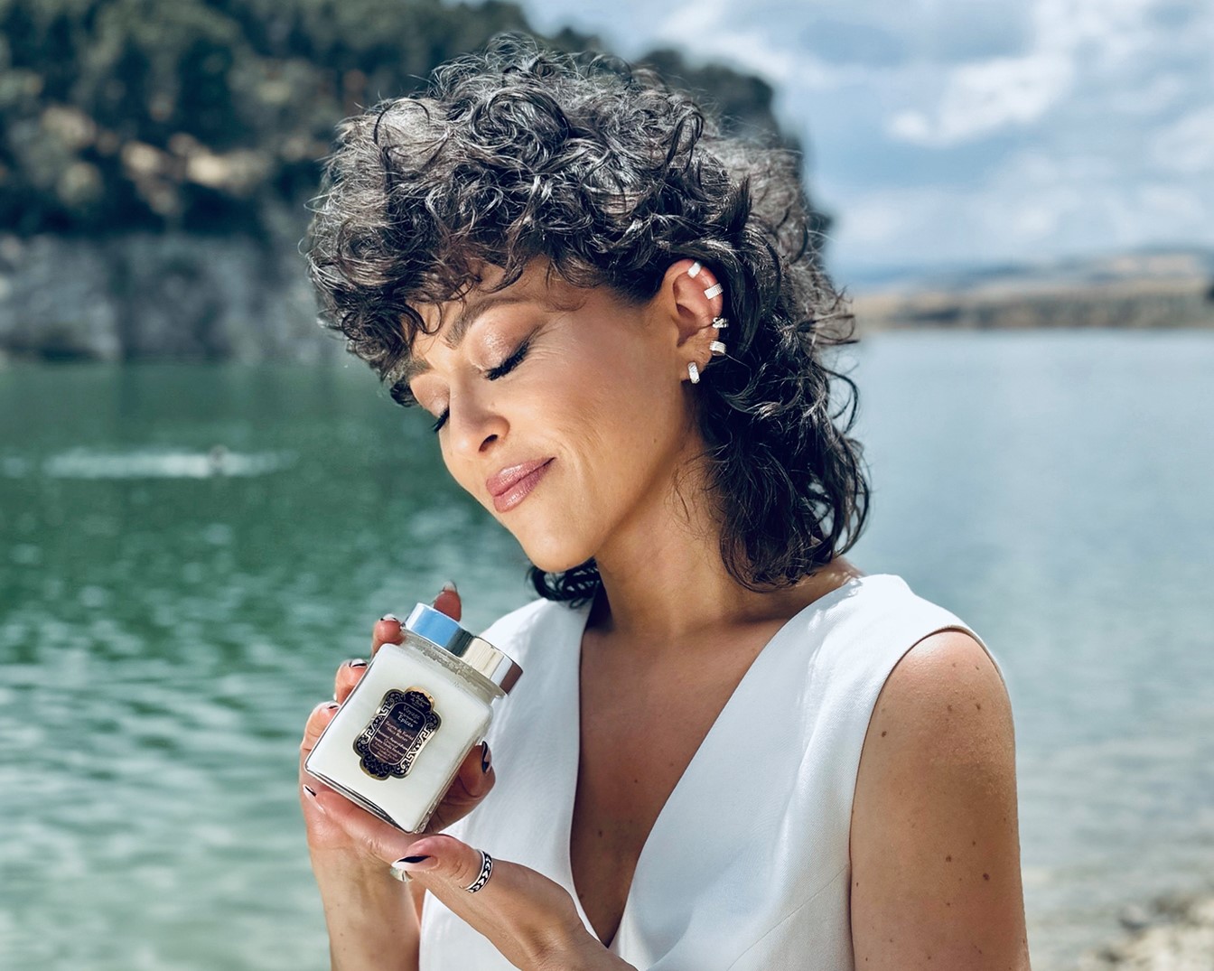 Kobieta z kręconymi włosami w białej sukience trzyma w dłoniach świecę zapachową w szklanym słoiku z srebrną nakrętką, zamykając oczy i uśmiechając się, na tle jeziora.