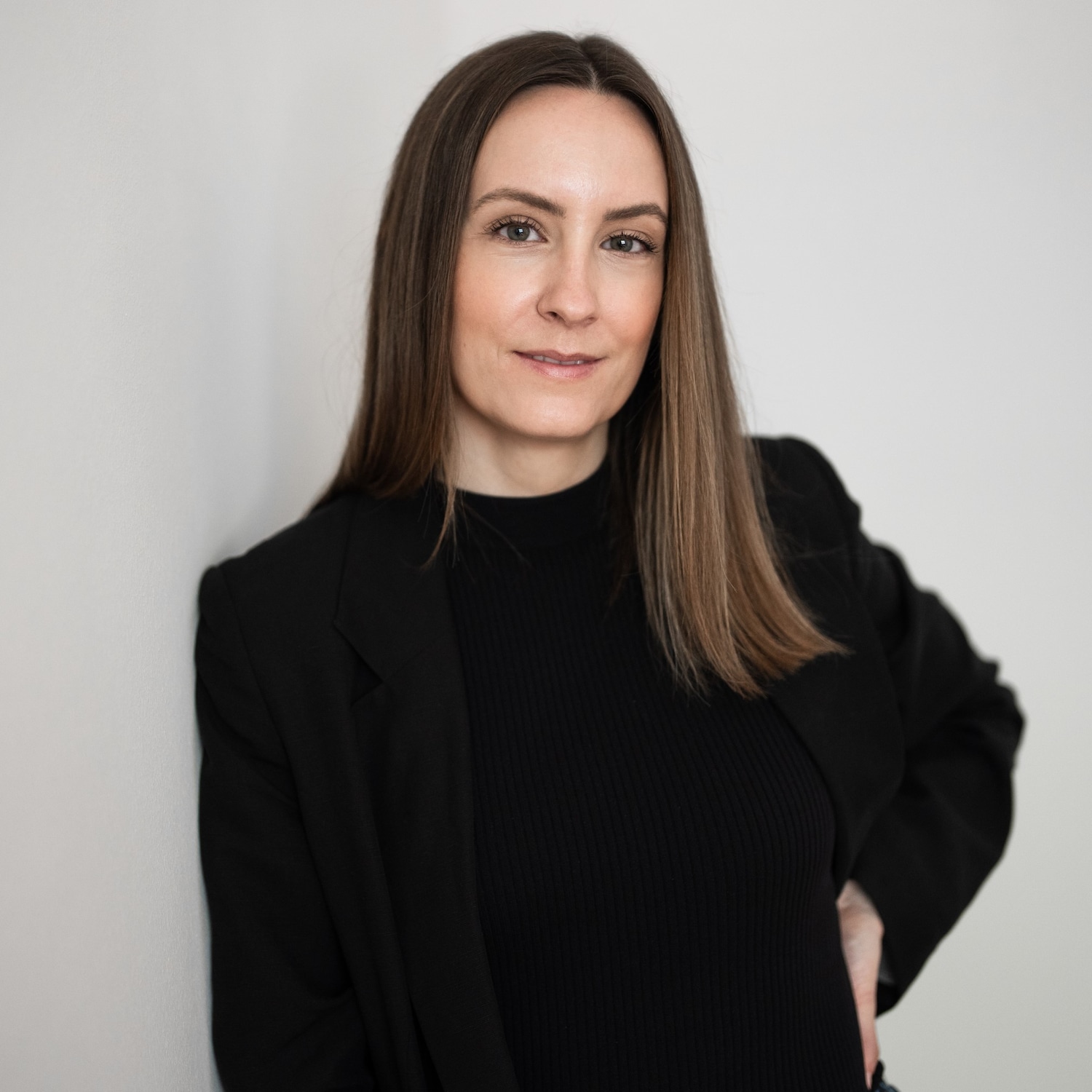 Portrait d'une femme aux cheveux longs et châtains, portant un blazer noir élégant sur un pull noir à col montant, posant avec assurance devant un mur blanc.
