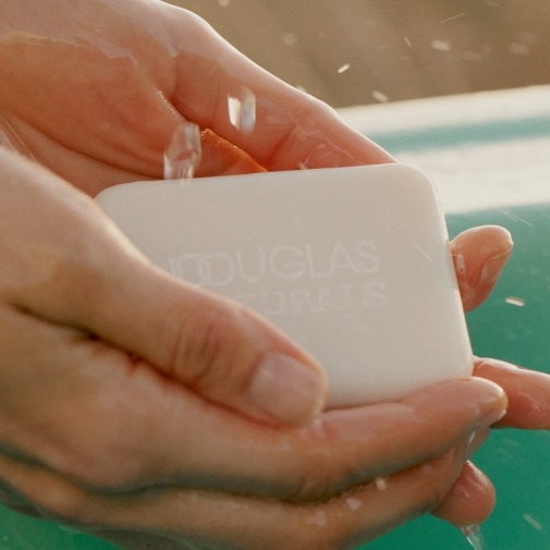 Dłonie trzymają białe mydło w kostce Douglas Naturals pod strumieniem wody, co sugeruje naturalne i czyste składniki.