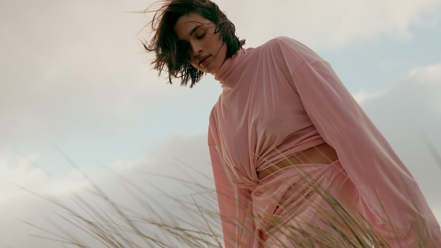 Kobieta o krótkich, ciemnych włosach ubrana w różowy komplet z golfem, pozuje na tle nieba.