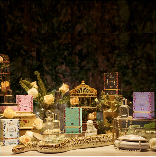 Esplora Jardin Bohème, una fragranza in flaconi colorati e confezioni decorate con motivi floreali, esposta tra rose, gabbie dorate e una scultura di serpente.