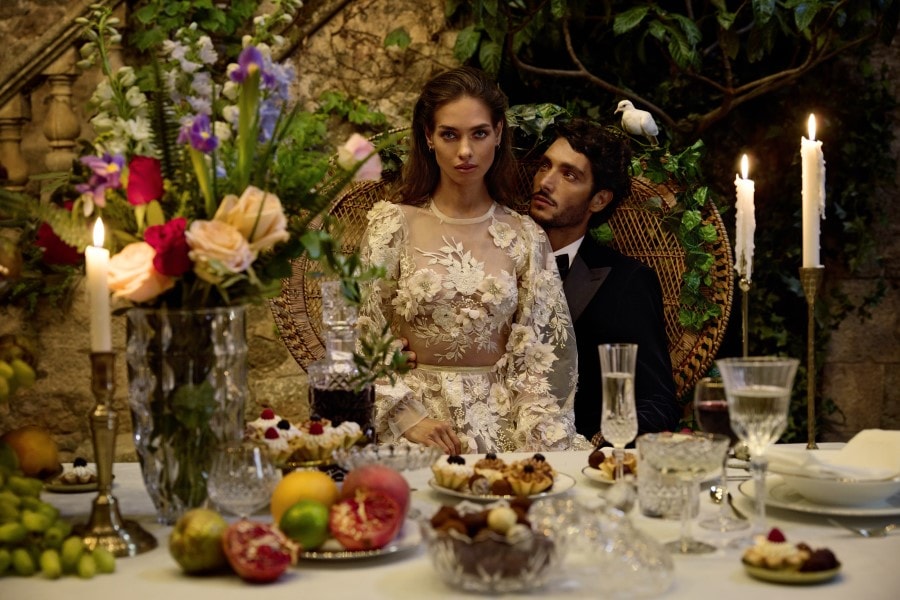 Un couple élégant est assis à une table somptueuse, entouré de fleurs, de bougies et de desserts, créant une ambiance romantique et intime.