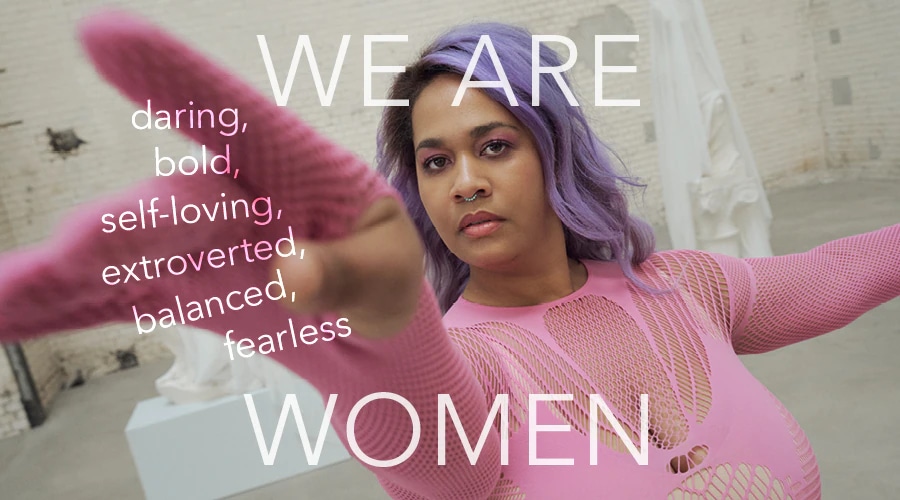 A woman with purple hair, wearing a pink fishnet top, confidently extends her arm towards the camera, embodying the words 'We are women: daring, bold, self-loving, extroverted, balanced, fearless'.