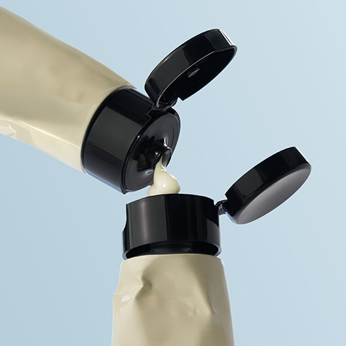 Nahaufnahme von zwei beigen Tuben mit schwarzen Klappdeckeln, aus denen eine helle Creme austritt.