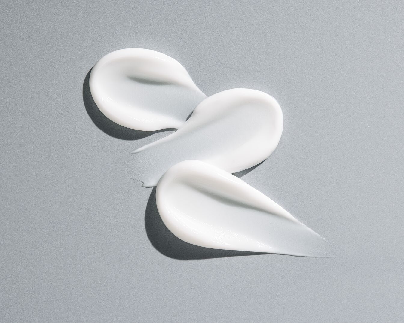 Tres trazos de crema blanca y brillante sobre un fondo gris claro, mostrando una textura rica e hidratante.