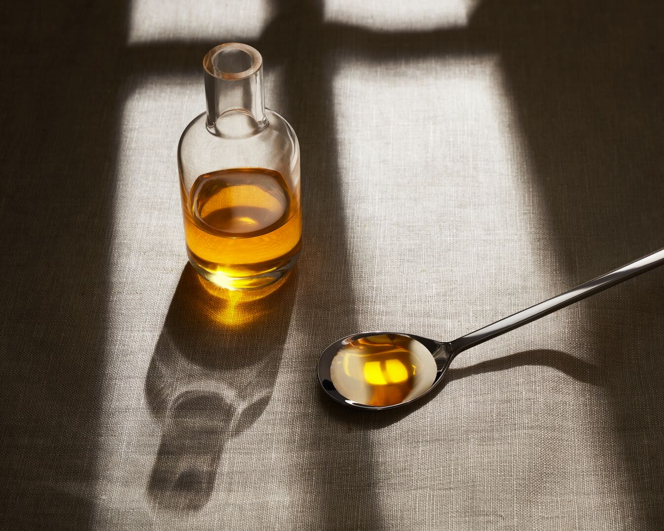 Aceite dorado en una botella de vidrio transparente y una cuchara de metal sobre un mantel texturizado, capturado bajo una luz brillante.