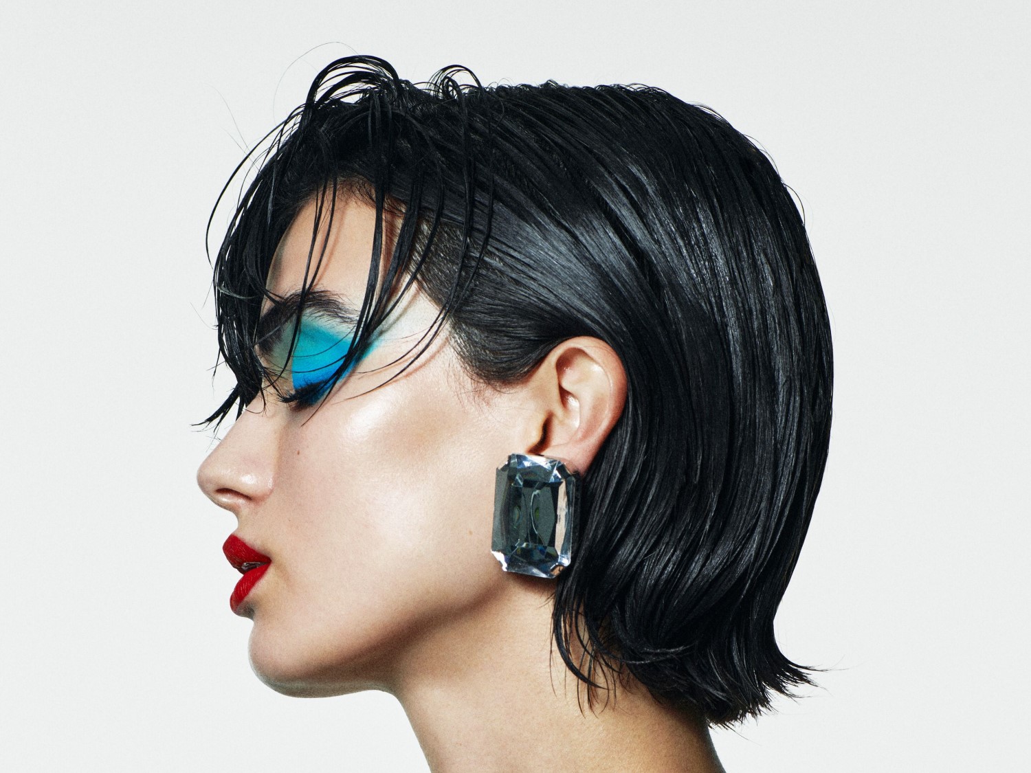 Profil einer Frau mit glänzendem, kurzem, schwarzem Haar, blauem Lidschatten, rotem Lippenstift und einem großen, quadratischen Ohrring.