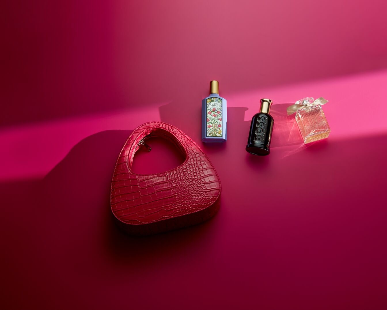 Auf einem pinkfarbenen Hintergrund sind eine rote Handtasche mit Krokoprägung, ein blassblaues Parfüm mit Blumenmuster, ein schwarzer Boss-Duft und ein durchsichtiges Parfüm mit Schleife zu sehen.