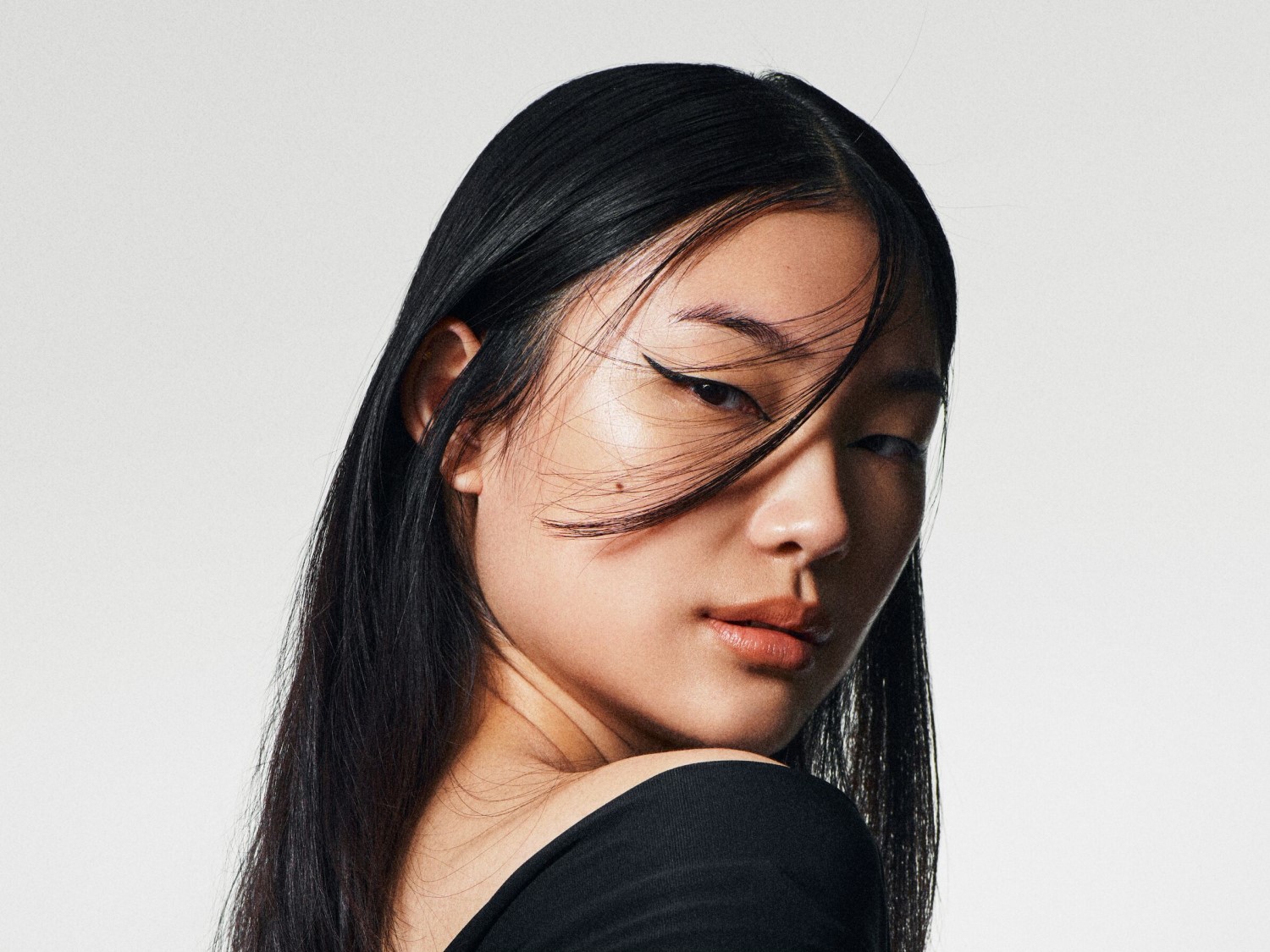 Een close-up van een vrouw met lang, zwart haar en een strakke eyeliner, die over haar schouder kijkt met een intense blik.