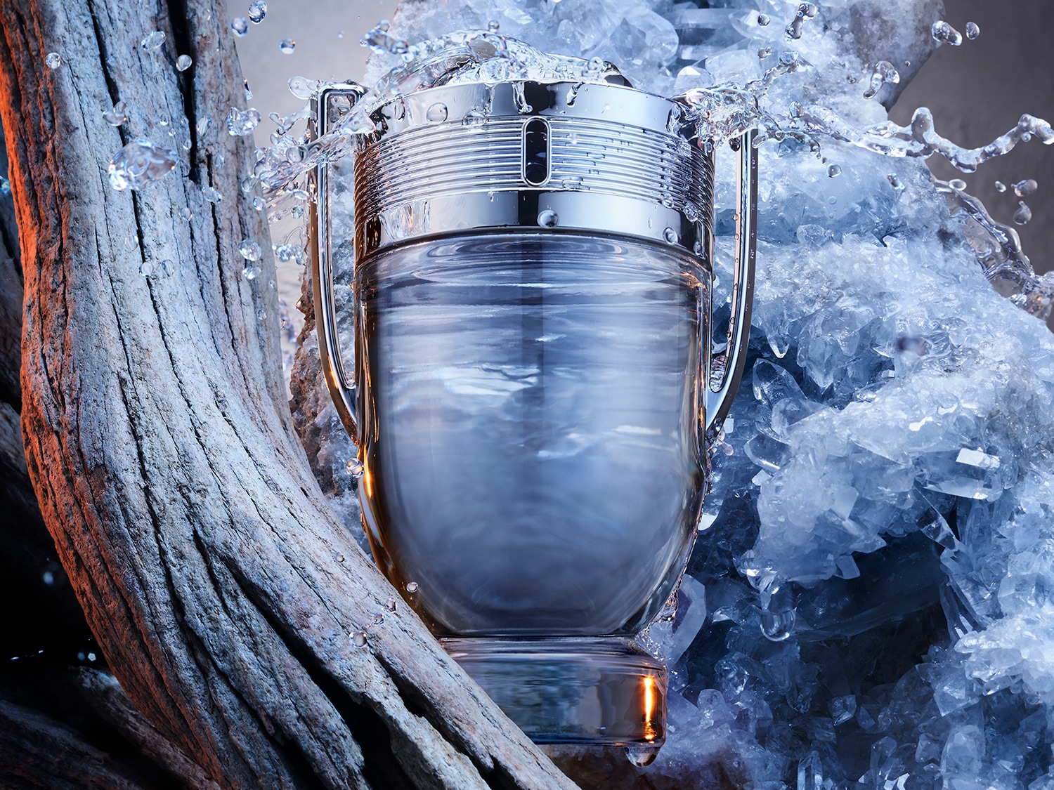 El frasco de perfume con forma de trofeo de plata está rodeado de salpicaduras de agua, colocado sobre un lecho de cristales azules y un tronco de árbol.