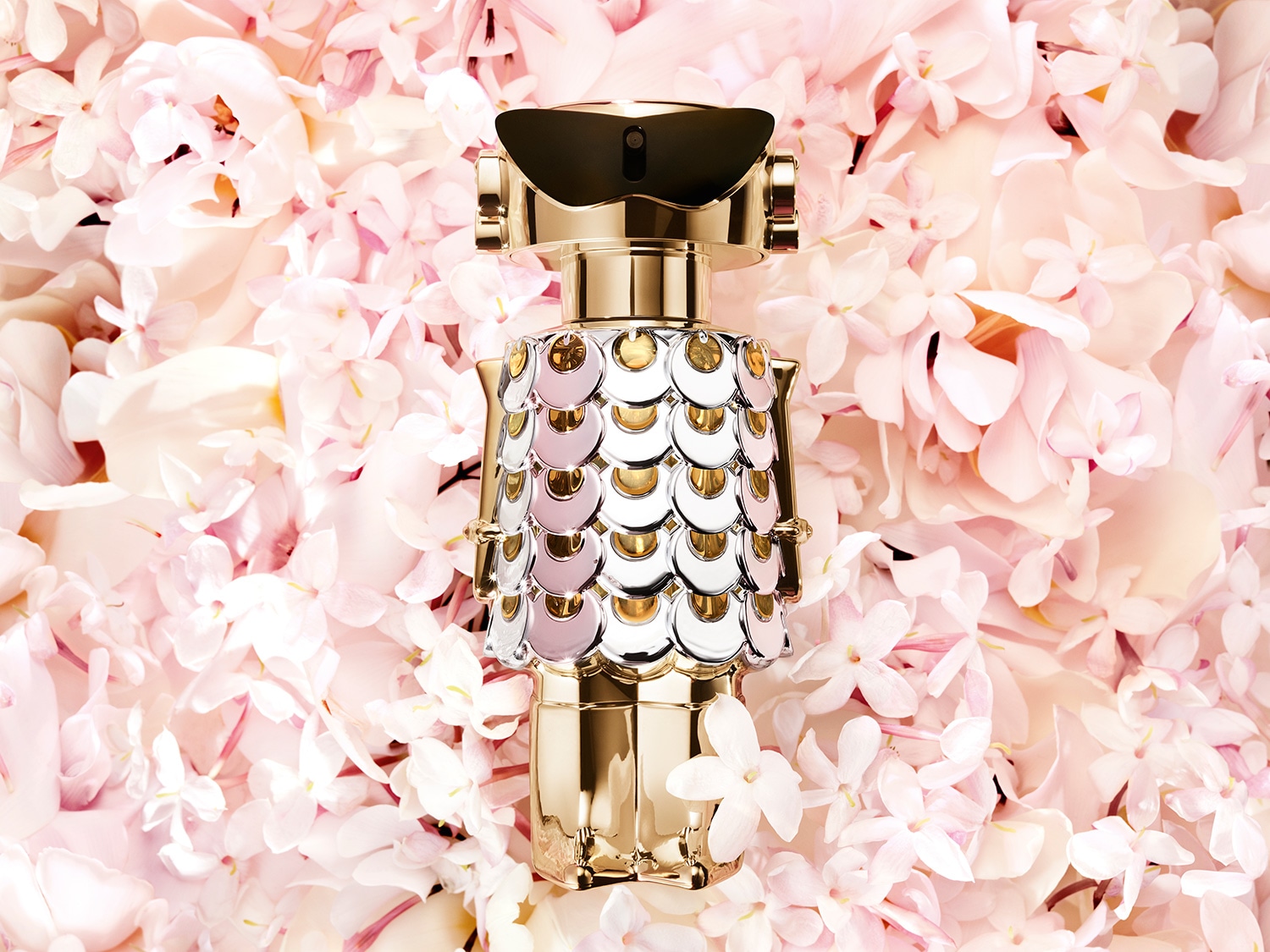 El frasco dorado de perfume con detalles en forma de escamas plateadas y rosadas destaca sobre un lecho de delicadas flores rosadas, evocando lujo y feminidad.