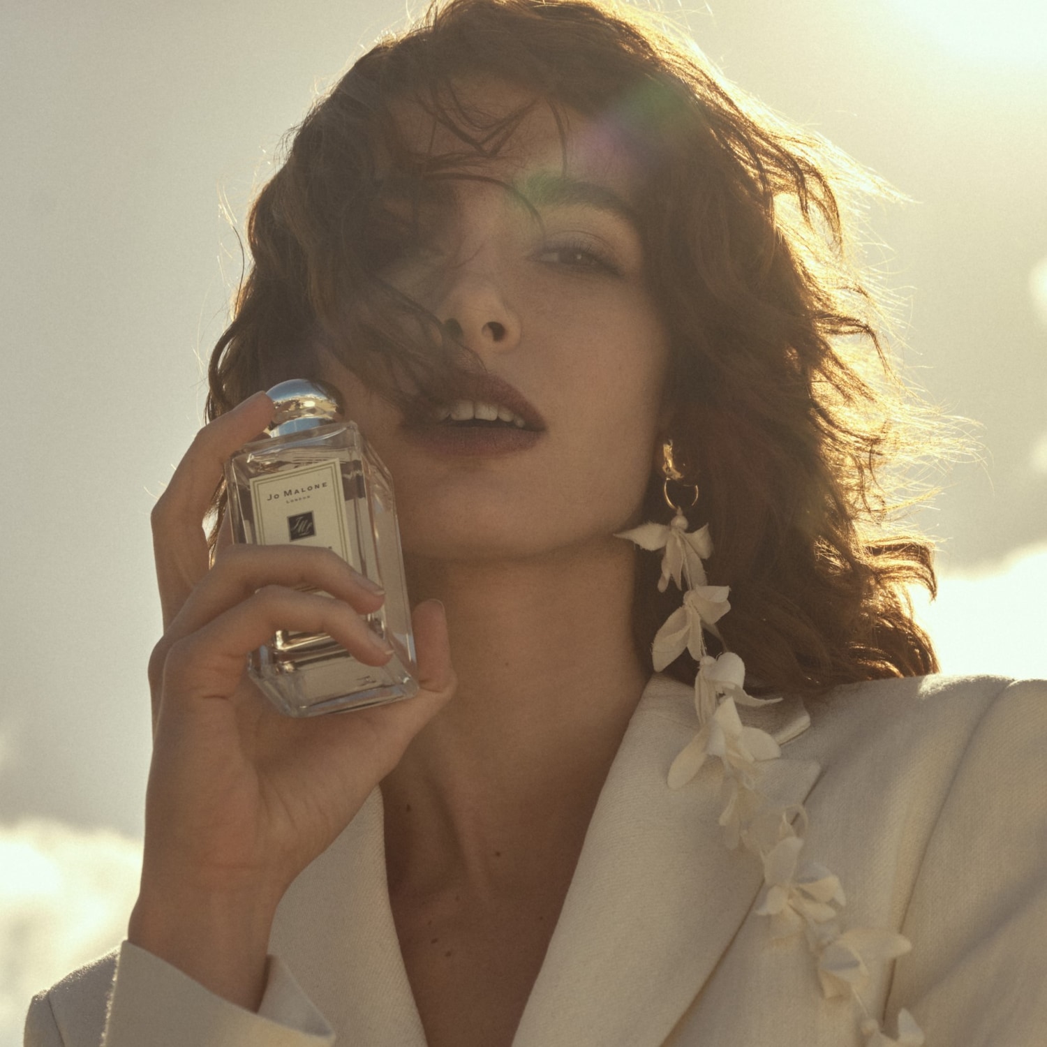 Eine Frau mit gewelltem Haar hält eine durchsichtige Flasche Jo Malone Parfüm vor einem sonnigen Himmel.