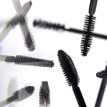 Na białym tle widoczne są czarne szczoteczki do tuszu do rzęs, tworzące dynamiczny i abstrakcyjny wzór.