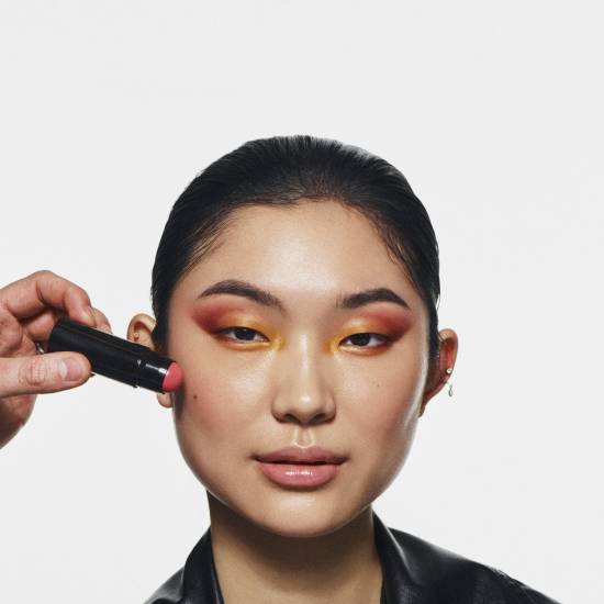 Na twarzy kobiety z makijażem oczu w odcieniach żółci i czerwieni nakładany jest róż w sztyfcie o kremowej konsystencji.