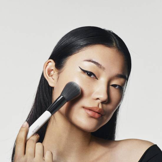 Portret kobiety z czarnymi włosami i eyelinerem, która nakłada puder pędzlem z białym trzonkiem.